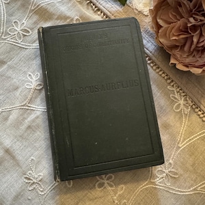 May include: A dark green book titled "Bevan's Origins of Christianity" and "Marcus Aurelius". The book is rectangular and appears vintage, with a slightly worn cover. A pink flower and greenery are in the background.