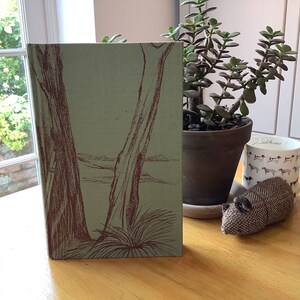 May include: A hardcover book with a light green cover featuring a brown illustration of trees and foliage. The book is standing upright on a wooden surface, next to a potted plant, a mug with dog illustrations, and a fabric mouse.