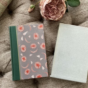 Puede incluir: Un libro de tapa dura con una cubierta gris con un patrón de formas circulares y triangulares rojas y blancas. El lomo es verde con un borde dorado. Un libro abierto con una cubierta gris claro está al lado. Una rosa rosa y un cojín están en el fondo.