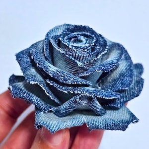 Könnte beinhalten: Eine Rose aus blauem Denim. Die Blume besteht aus recyceltem Denim-Stoff mit detaillierter Blütenblattkonstruktion. Die Ränder der Blütenblätter sind ausgefranst und verleihen Textur. Ein einzigartiger, umweltfreundlicher Dekorationsartikel.