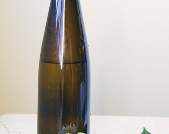 May include: A dark brown glass bottle with a clear liquid inside, sitting on a white surface with white flowers and green leaves.