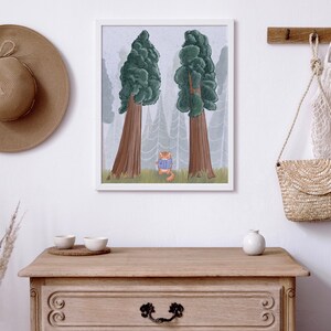 May include: A framed illustration of a cat wearing a blue sweater standing in a foggy forest with two large trees. The illustration is hanging on a white wall above a wooden dresser with a straw hat, a woven bag, and a ceramic vase.
