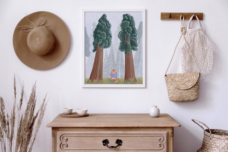 May include: A framed illustration of a cat wearing a blue sweater standing in a foggy forest with two large trees. The illustration is hanging on a white wall above a wooden dresser with a straw hat, a woven bag, and a ceramic vase.