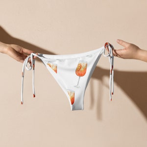 May include: White bikini bottom with a tie-side design. The fabric features a repeating pattern of orange and yellow cocktails.