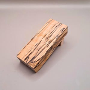 May include: A wooden block with a natural wood finish and dark brown streaks. The block is shaped like a small, slanted platform.