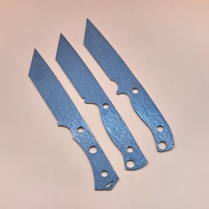 May include: Three metal knife blanks with rounded tips and holes for handles. The blanks are a light grey colour and have a textured surface.