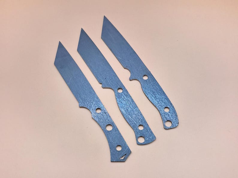 May include: Three metal knife blanks with rounded tips and holes for handles. The blanks are a light grey colour and have a textured surface.