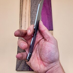 May include: A close-up of a silver knife blade with a black handle. The knife is held in a hand and is resting on a wooden surface with a purple and brown wood grain.