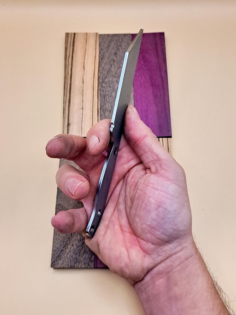 May include: A close-up of a silver knife blade with a black handle. The knife is held in a hand and is resting on a wooden surface with a purple and brown wood grain.
