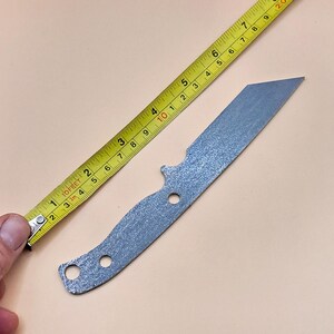 May include: A silver knife blank with a textured finish, measuring approximately 18 cm in length. The knife blank is being measured with a yellow tape measure.