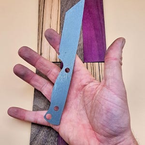May include: A silver knife blank with two holes in the handle. The blank is sitting on a piece of wood with different coloured wood grain.