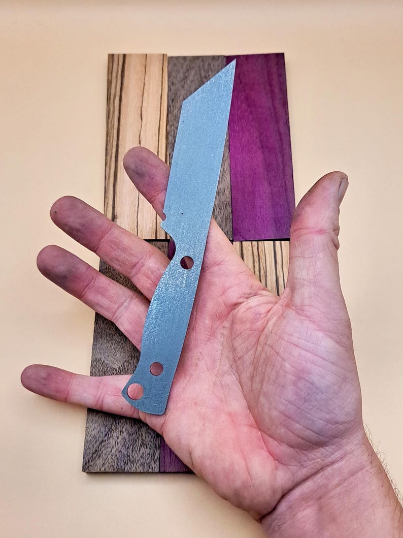 May include: A silver knife blank with two holes in the handle. The blank is sitting on a piece of wood with different coloured wood grain.
