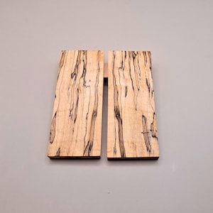 Op de afbeelding: Twee rechthoekige houten planken met een lichtbruine basis en donkerbruine adering. De planken worden samengevoegd door een dun, lichtgekleurd houten stuk. Het houtnerfpatroon is zichtbaar. De achtergrond is effen grijs.