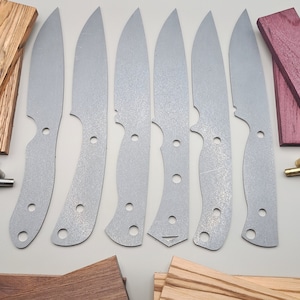 May include: A set of six knife blanks, made of metal, with a silver finish. The blanks are arranged in a row on a white surface. There are also pieces of wood in various colors and sizes, as well as two brass tubes.