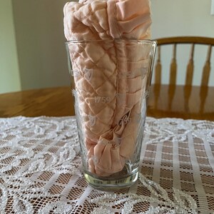 May include: A clear glass with the year 1759 printed on it, holding a folded, quilted, peach-colored fabric. The glass sits on a white lace tablecloth, with a wooden table and a chair in the background.