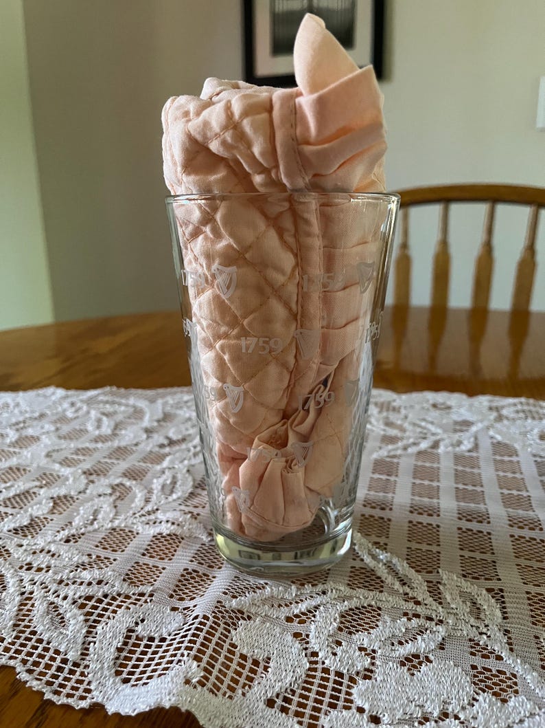 May include: A clear glass with the year 1759 printed on it, holding a folded, quilted, peach-colored fabric. The glass sits on a white lace tablecloth, with a wooden table and a chair in the background.