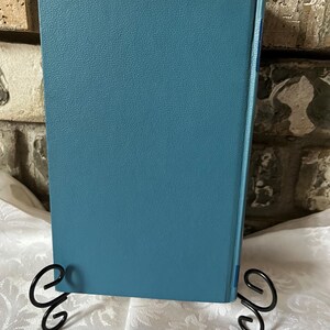 May include: A teal blue hardcover book with a textured cover. The book is standing upright on a white surface with two black metal bookends.