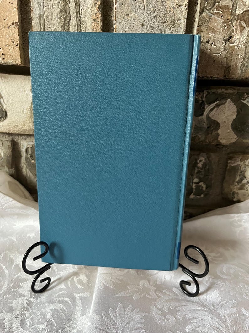 May include: A teal blue hardcover book with a textured cover. The book is standing upright on a white surface with two black metal bookends.