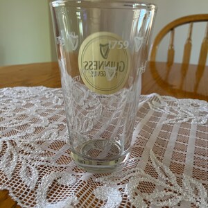 May include: A clear pint glass featuring the Guinness logo and the year 1759. The glass is placed on a white lace tablecloth. The glass is empty and clean.