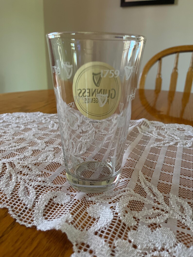 May include: A clear pint glass featuring the Guinness logo and the year 1759. The glass is placed on a white lace tablecloth. The glass is empty and clean.
