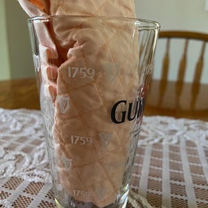 May include: A clear pint glass with the Guinness logo and the year 1759 printed on it. Inside the glass is a peach-colored, quilted fabric. The glass is sitting on a white lace tablecloth.