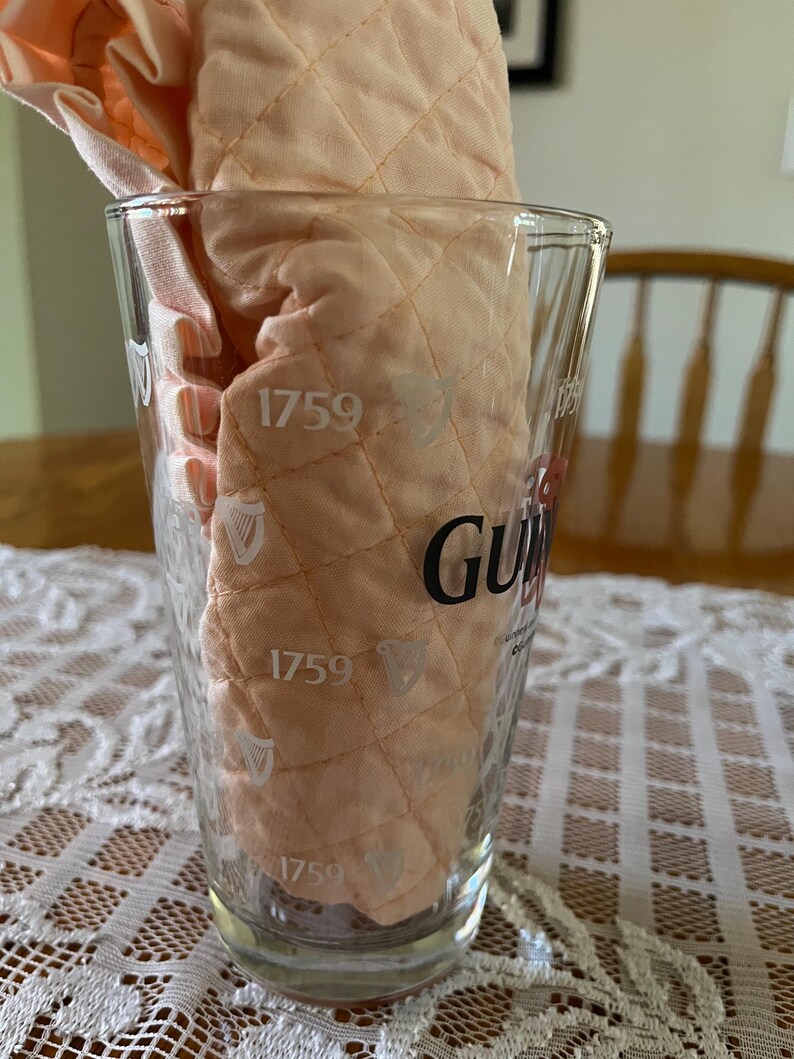 May include: A clear pint glass with the Guinness logo and the year 1759 printed on it. Inside the glass is a peach-colored, quilted fabric. The glass is sitting on a white lace tablecloth.