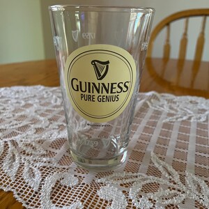 May include: A clear pint glass with the Guinness logo, featuring a harp and the words "Guinness Pure Genius" on a cream-coloured oval. The glass also has the year "1759" printed on it. The glass is on a white lace tablecloth.