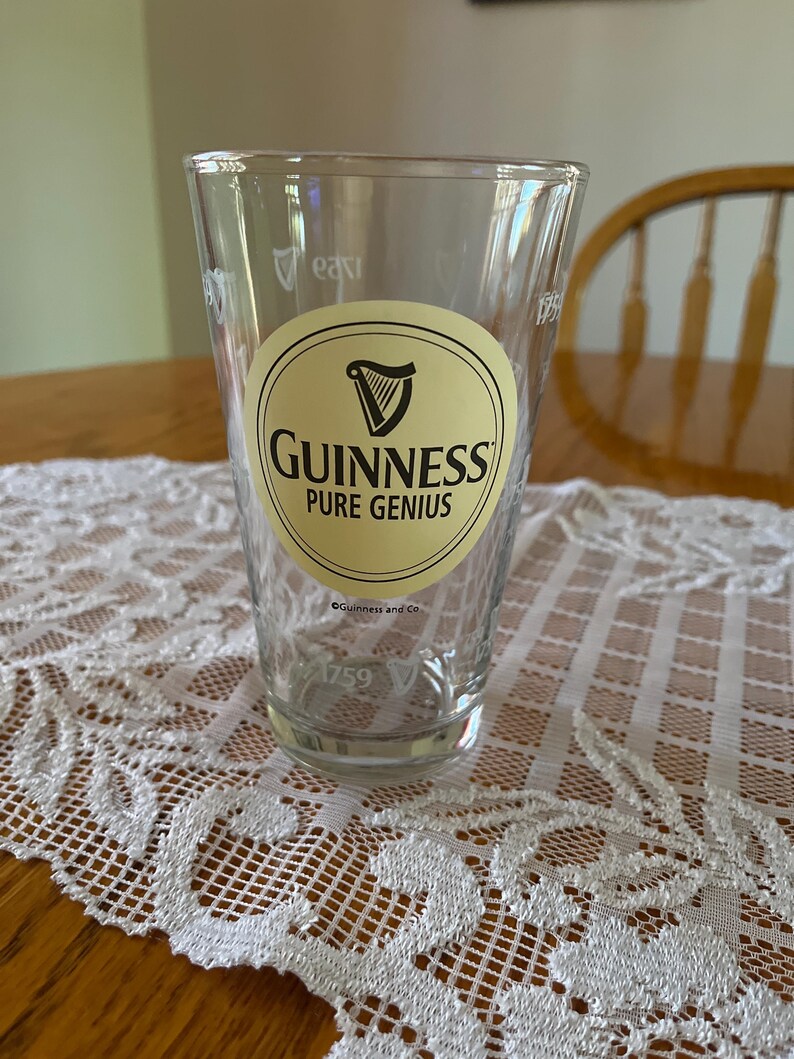 May include: A clear pint glass with the Guinness logo, featuring a harp and the words "Guinness Pure Genius" on a cream-coloured oval. The glass also has the year "1759" printed on it. The glass is on a white lace tablecloth.