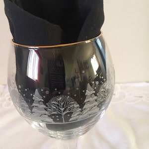 May include: A clear glass wine glass with a gold rim and a white etched design of a winter scene with trees and snowflakes. The glass is partially covered by a black cloth napkin.