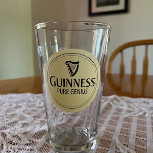 May include: A clear pint glass with the Guinness logo, featuring a harp within a cream-coloured oval and the words "Guinness Pure Genius." The glass has a slightly tapered shape and is set on a white lace tablecloth.