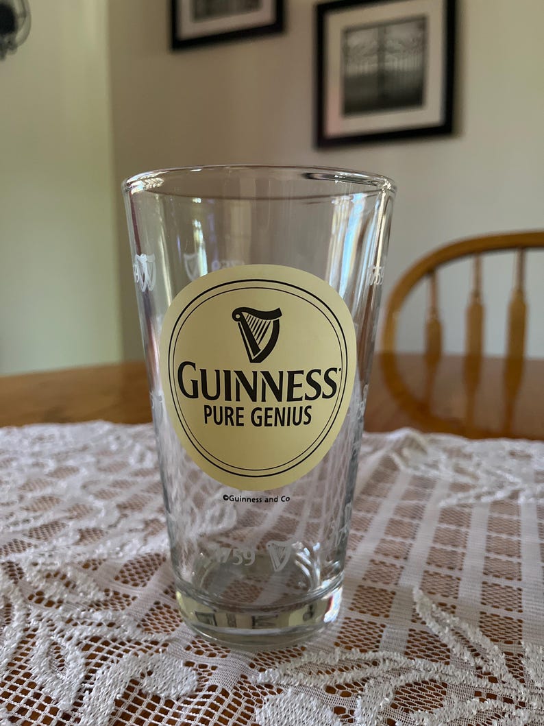 May include: A clear pint glass with the Guinness logo, featuring a harp within a cream-coloured oval and the words "Guinness Pure Genius." The glass has a slightly tapered shape and is set on a white lace tablecloth.