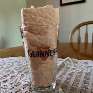 May include: A clear pint glass with the Guinness logo and the year 1759 printed on it. The glass is filled with a light peach-colored, quilted fabric. The glass sits on a white lace tablecloth.
