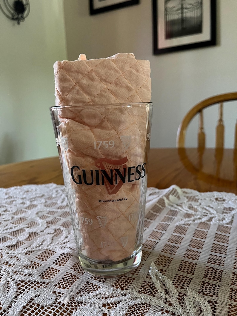 May include: A clear pint glass with the Guinness logo and the year 1759 printed on it. The glass is filled with a light peach-colored, quilted fabric. The glass sits on a white lace tablecloth.