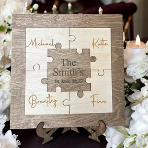 May include: A wooden puzzle-shaped sign with a light brown frame. The centre piece reads "The Smith's Est. October 11th, 2025." Individual puzzle pieces are engraved with names: Michael, Katie, Brantley, and Finn. The sign is displayed on a wooden stand.