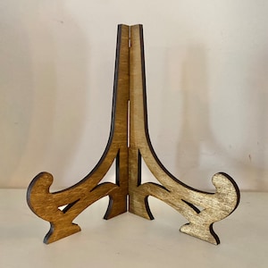 May include: A wooden display stand with a natural finish. The stand has a three-legged base with decorative scrollwork and a tall, vertical support. The wood grain is visible, and the stand is designed to hold a picture or plate.