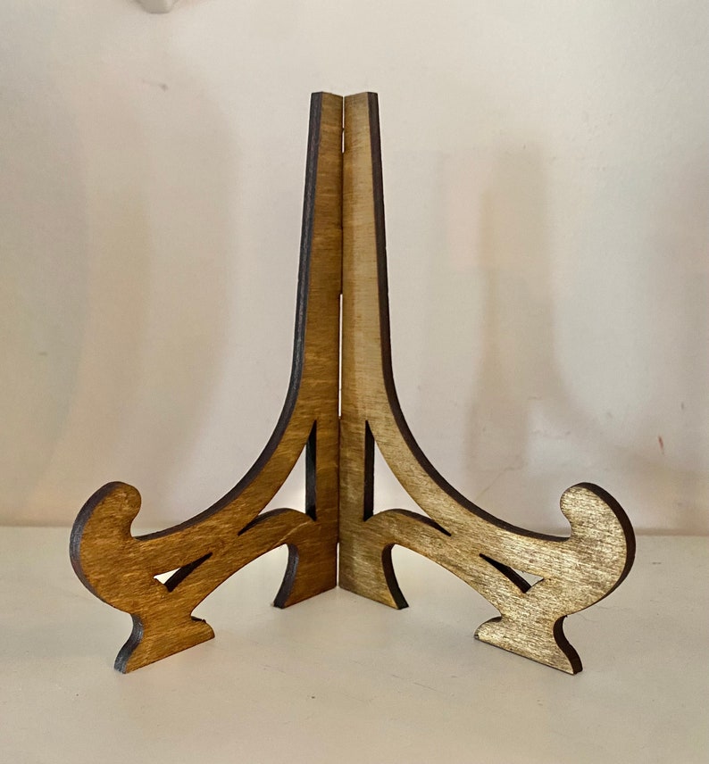 May include: A wooden display stand with a natural finish. The stand has a three-legged base with decorative scrollwork and a tall, vertical support. The wood grain is visible, and the stand is designed to hold a picture or plate.
