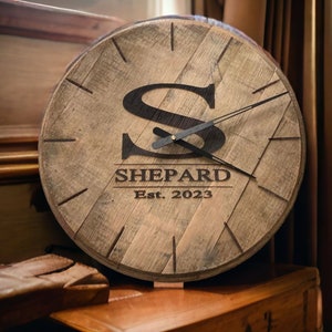 Può includere: Un orologio in legno con una lancetta delle ore e dei minuti in metallo nero. Il quadrante dell'orologio ha una grande lettera nera "S" e il testo "SHEPARD Est. 2023".