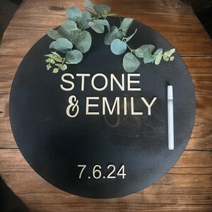 Peut inclure: Panneau rond en bois noir avec les noms "STONE & EMILY" et la date "7.6.24" en couleur claire. Le panneau est orné de feuilles d'eucalyptus vertes et d'un marqueur blanc.