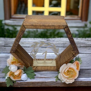 May include: A wooden hexagonal table number holder with a laser-cut number 1 and floral accents. The holder is made of dark brown wood and features a clear acrylic insert with a laurel wreath design. Peach and white flowers are attached to the base.