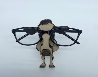 May include: A wooden cow figurine with black and white markings wearing black framed glasses. The cow is standing on a white surface.