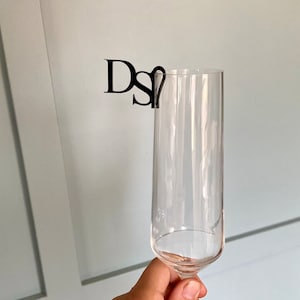 May include: A clear glass champagne flute with a black plastic monogrammed charm attached to the rim. The charm reads "DS".