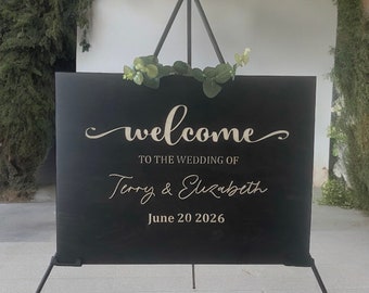 May include: Black welcome sign with gold lettering. The sign reads "Welcome to the wedding of Terry & Elizabeth June 20 2026".