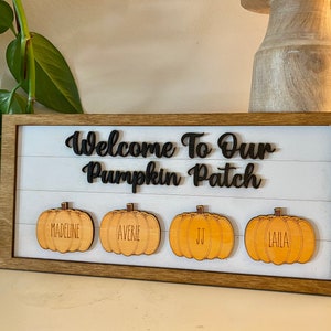 Op de afbeelding: Welkom bij onze pompoen patch bord met een houten frame en vier houten pompoenen met de namen Madeline, Averie, JJ en Laila.