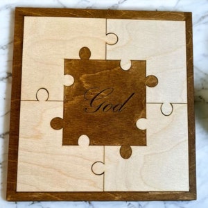 May include: A wooden jigsaw puzzle featuring the word "God" engraved in the centre. The puzzle is constructed from light and dark wood pieces, with a dark brown frame. The pieces are interlocked, forming a square.