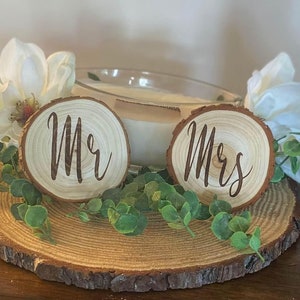 May include: Two wooden slices with the words "Mr" and "Mrs" engraved on them. The slices are placed on a larger wooden slice with greenery and white flowers around them.