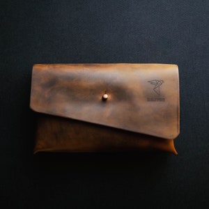 May include: A brown leather clutch with a flap closure and a small metal button. The clutch has a logo that says "The Paper Plane" and "Handmade in Italy".