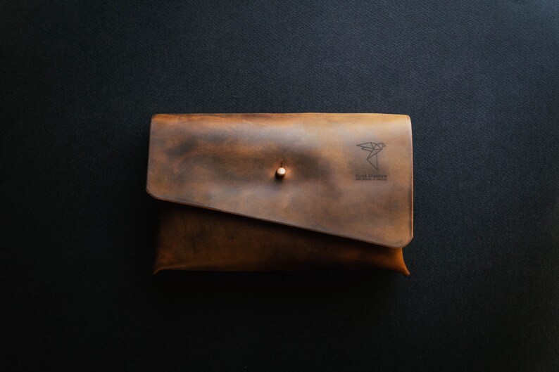 May include: A brown leather clutch with a flap closure and a small metal button. The clutch has a logo that says "The Paper Plane" and "Handmade in Italy".