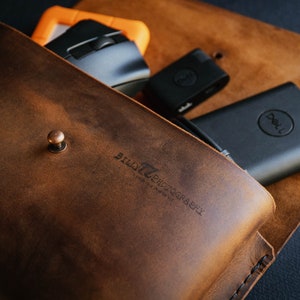 May include: A brown leather pouch with a snap closure and the text "BILLY PHOTOGRAPHY" embossed on the front. The pouch is open and contains a black computer mouse, a black USB drive, and a black Dell power bank.
