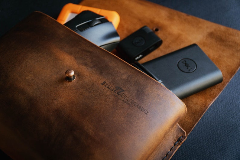 May include: A brown leather pouch with a snap closure and the text "BILLY PHOTOGRAPHY" embossed on the front. The pouch is open and contains a black computer mouse, a black USB drive, and a black Dell power bank.