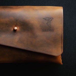 May include: A brown leather clutch with a brass button closure. The clutch has a geometric bird design and the text "Olive Sparrow Handmade in Greece" embossed on the front.